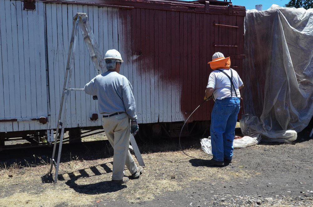 2018-06-26 With the end of an already completed car covered, the paint crew starts spriaying box car red on the preped car.jpg